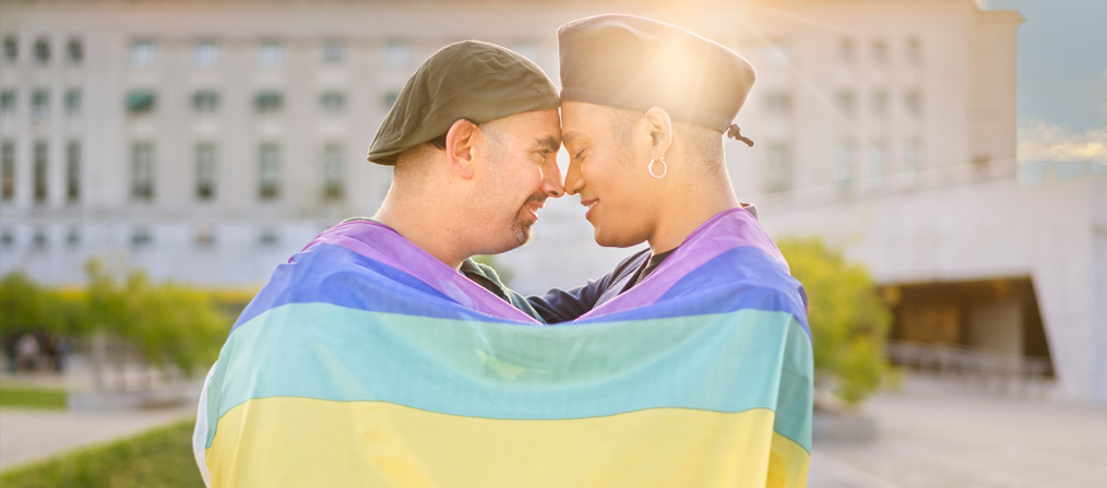 Dos personas envueltas en la bandera LGBTIQ+, juntando las frentes y sonriendo en una muestra de amor